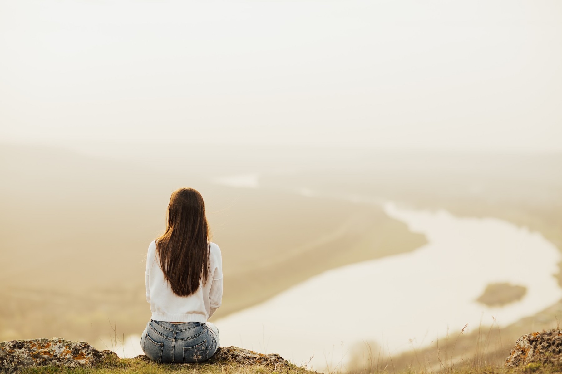 Back view of woman sitting on top of the hill and looking at river in evening sunset young traveler