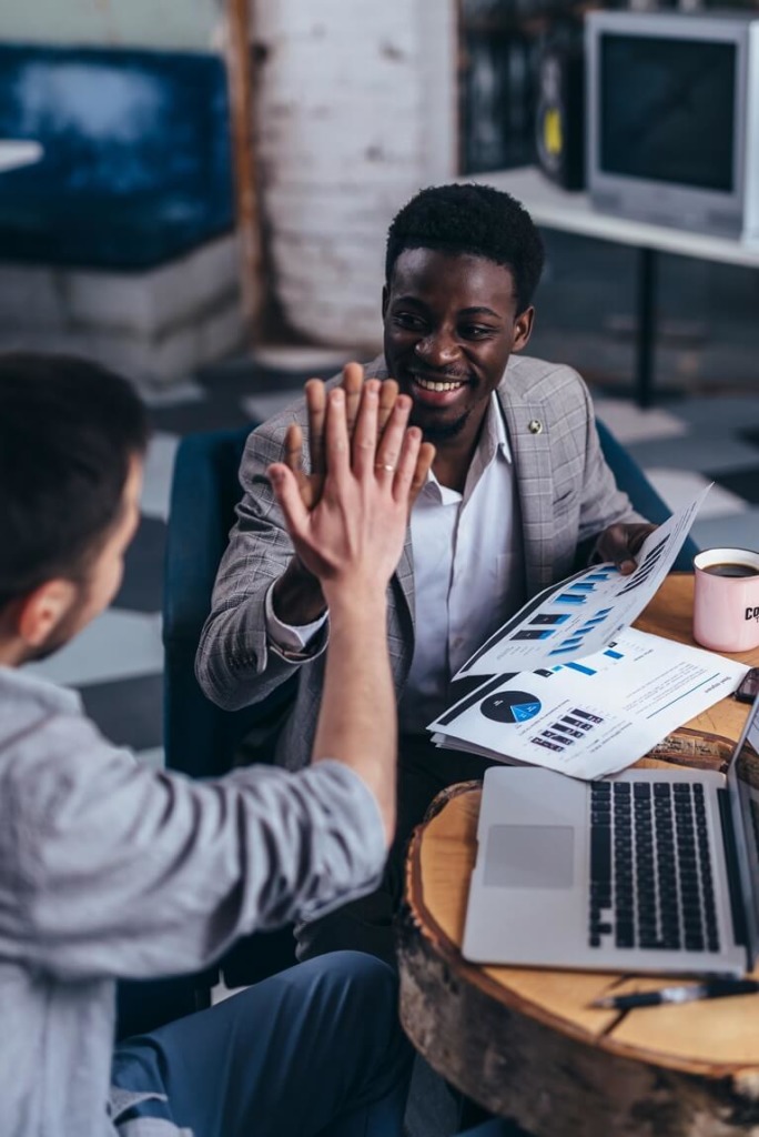 African American male business partners giving high five at table to Caucasian coworker