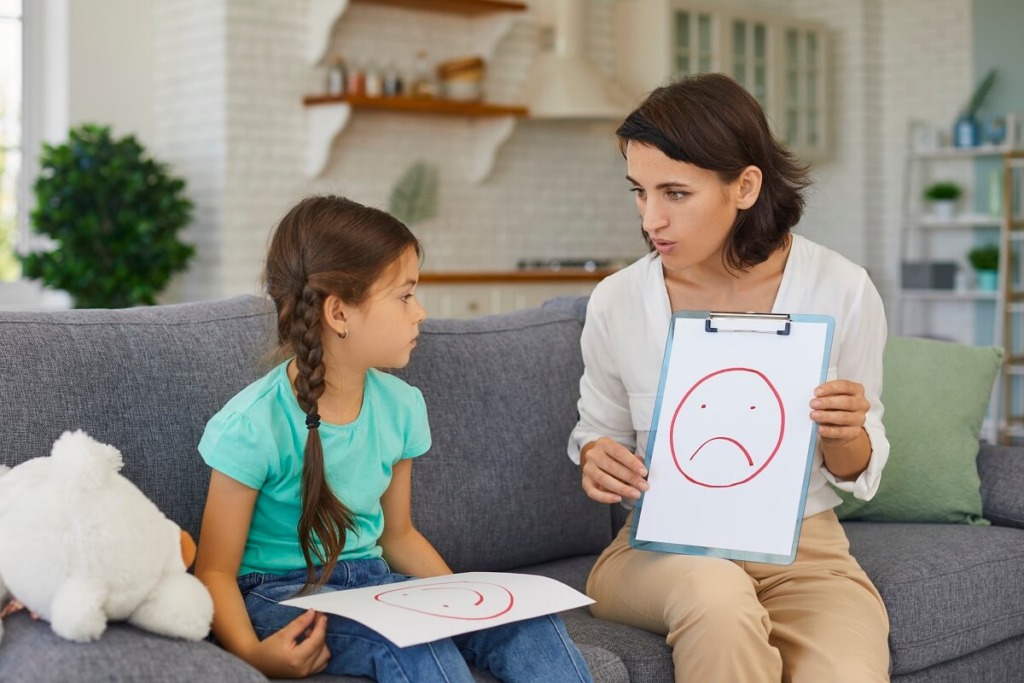 Mother at home discussing emotions with daughter using various emotion faces drawn on paper