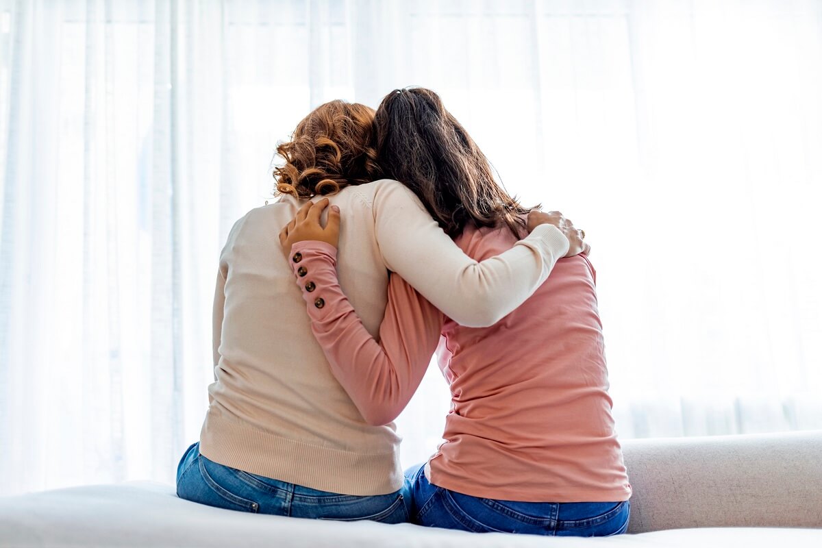 Rear back view mother and daughter embrace sitting on bed at home