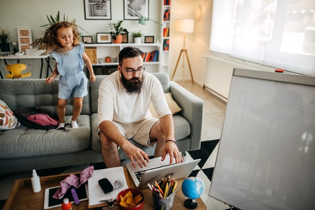 Bearded father works from home in living room while energetic neurodiverse daughter jumps on couch
