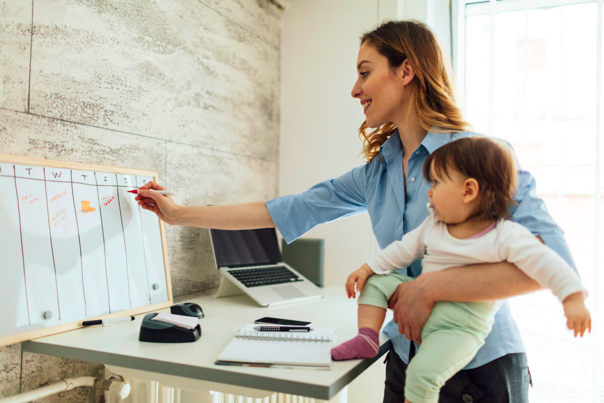 Mother working from home carrying her cute baby girl while writing on whiteboard calendar