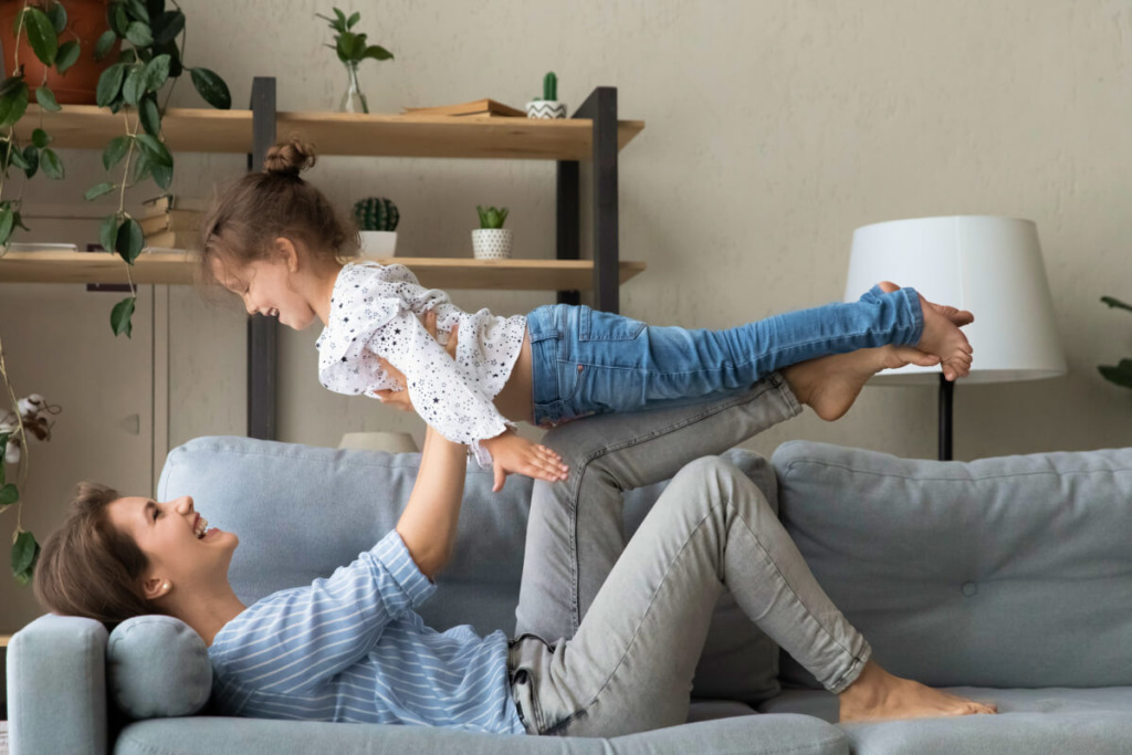 Happy mom lifting little daughter kid up in air, playing airplane with girl, lying on couch