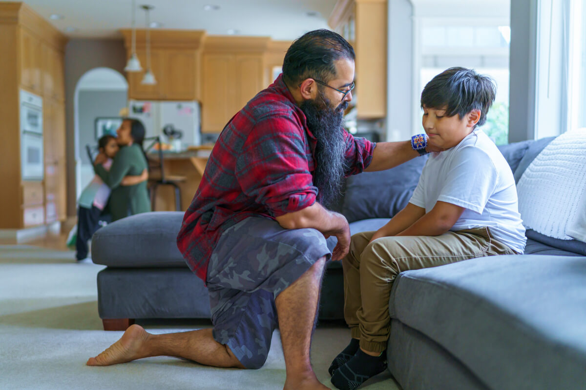 son sits on couch while dad has serious conversation, mother and daughter hug in background