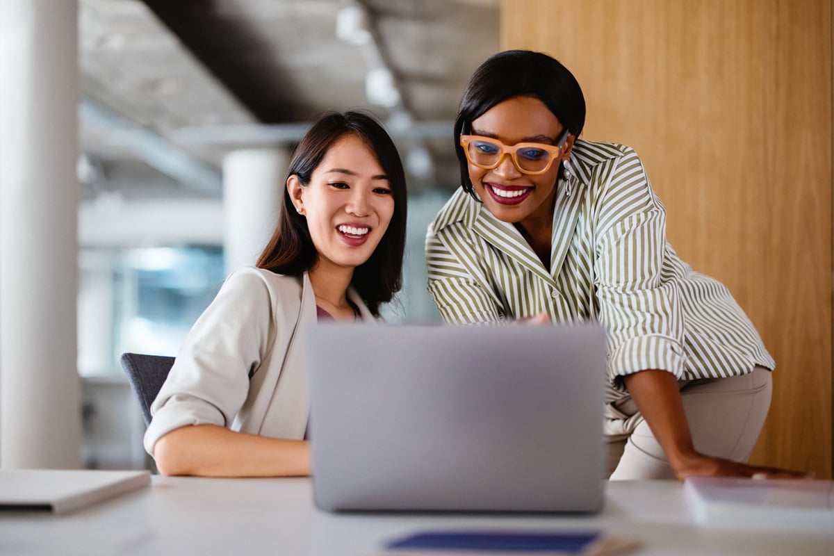 Two professional women in modern office discussing work on laptop