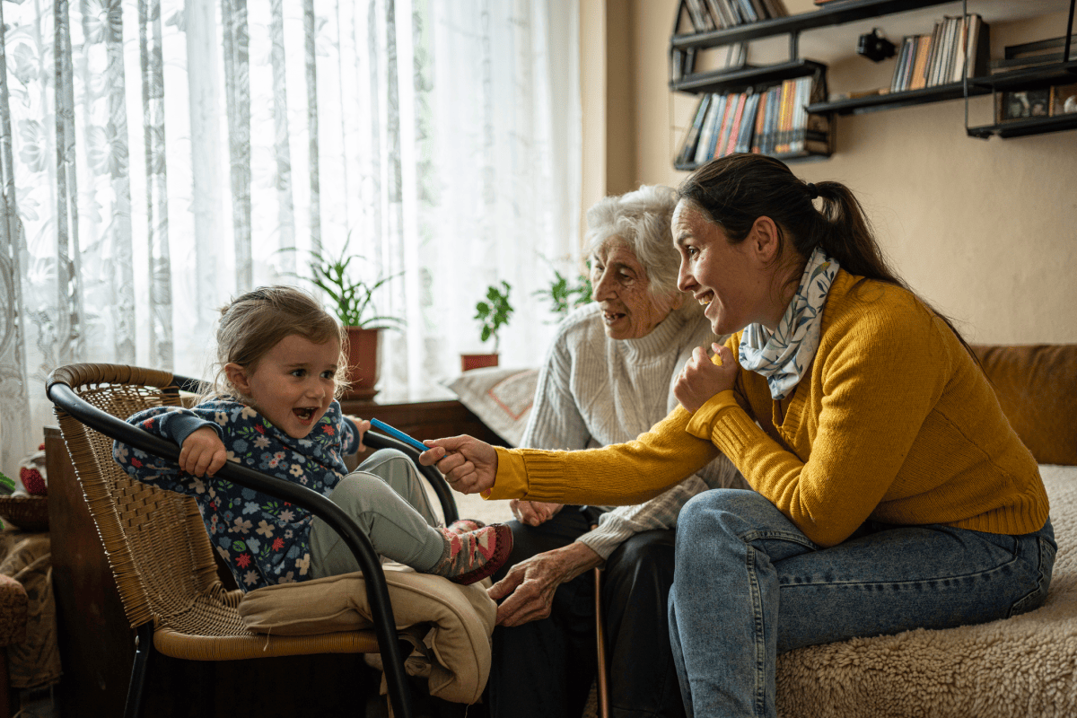 A child smiles while interacting with two older family members in a bright, cozy living room.
